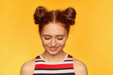 Teenage girl, happy looking, charming red hair woman with two buns. Wearing striped shirt and watching down with smile, shy. Closeup, stand isolated over yellow background