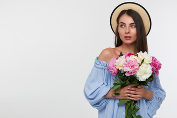 Young lady, pretty woman with long brunette hair. Wearing a hat and blue pretty dress. Holding a bouquet of beautiful flowers. Watching to the left at copy space over white background