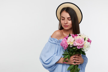 Teenage girl, happy looking woman with brunette long hair. Wearing a hat and blue dress. Holding a bouquet of beautiful flowers. Watching to the left lower corner at copy space over white background