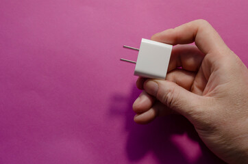 Man's hand holds white DC wall adapter against pink background.