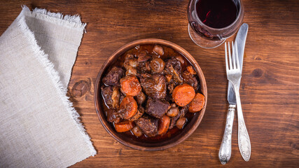 Ready burgundy beef in a clay bowl and a glass of red wine on a wooden table close-up, top view