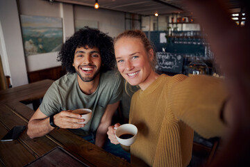 Portrait of cheerful young couple in modern cafe relaxing holding coffee mug and taking selfie