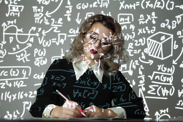 Intelligent young female maths student or teacher wearing glasses standing in front of a chalkboard with mathematical equations