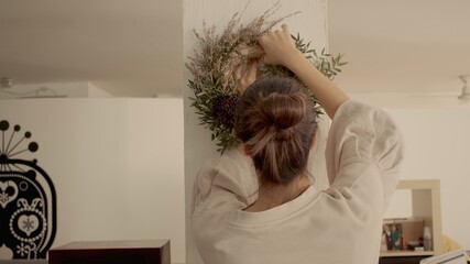 A young woman with brown hair in a bun in home wear is hanging a handmade winter festive wreath on the white column in her apartment. Light, warm atmosphere. Up view high-quality photo jpg.