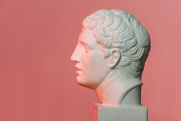 Profile view of a white marble head of young man over red-lit pink background. On a square pedestal.