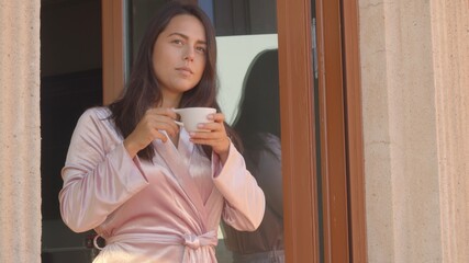Beautiful female with dark long hair in a pink silky bathrobe standing in the window of her house enjoying morning coffee from a white cup. Watching around. High-resolution medium shot portrait jpg