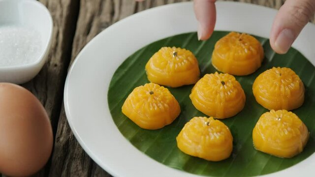 A woman's hand is delicately placing Thong-Eek, Sane Chan and Kanom Jamongkut (Thai sweets) on a white plate with banana leaves.