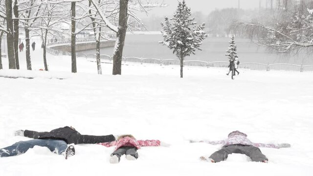 Family with kid making snowy angels in snow. Mother, father and daughter lying on the ground and moving like angels in snow in winter time. Family fun weekends with children near forest and lake 