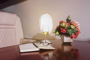 Flowers, diary, glass wine on work table of luxury interior in private jet. Modern and comfortable business airplane with decor. Concept of passenger service quality in aviation industry, at highest