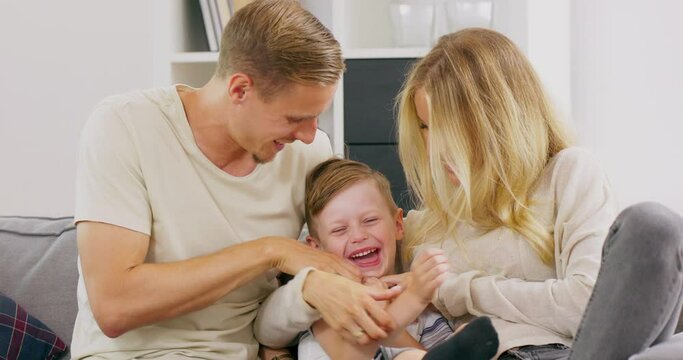 Affectionate caucasian father and mother tickling little adorable child son having fun laughing relaxing on sofa, polish parents playing with small kid boy bonding cuddling together at home.