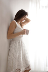 Middle age woman with white dress, drinking coffee at home, looking out of the window