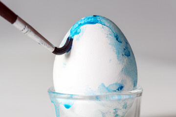 Brush is painting blue watercolor on an Easter egg for the holiday decoration, light grey background with copy space, close-up