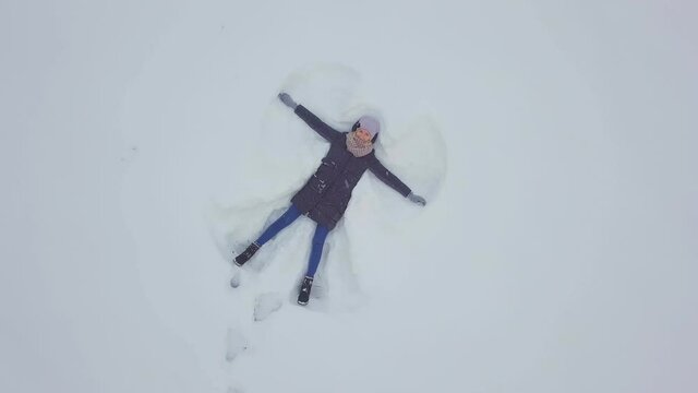 Aerial top view of happy Caucasian woman having fun during winter vacation, playing with snow, making Angel, carefree young woman feeling free during Christmas vacation