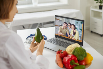 Woman doctor nutritiologist showing fresh ingredients and consulting young family online on laptop...