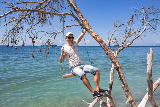 Summer landscape with a view of the sea. A guy sitting on a swing, on a fallen tree on the shore, and cargo ships on the horizon. The outskirts of the resort of Gelendzhik. Russia, Black Sea coast