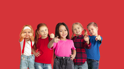 Pointing. Portrait of little caucasian children gesturing isolated on red studio background with copyspace. Cheerful kid models. Concept of human emotions, facial expression, sales, ad, childhood.