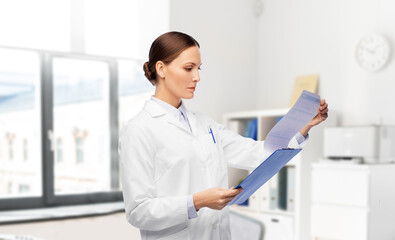 medicine, profession and healthcare concept - female doctor in white coat with clipboard reading medical report over hospital background