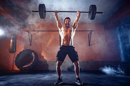 Muscular fitness man doing deadlift a barbell over his head in modern fitness center. Functional training. Snatch exercise. Smoke on background.