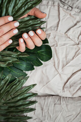 Womens hands with fresh neat white manicure in leaves of green plants, palm trees and monstera on background of crumpled beige fabric. Vertical content, selective focus