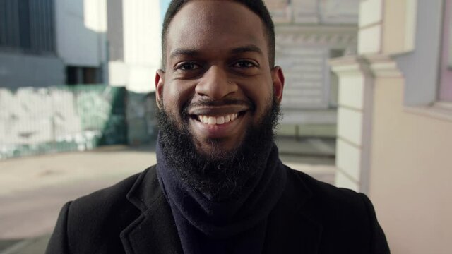 Close up portrait of cheerful bearded african american man smiling widely to camera outdoors, wearing scarf, slow motion
