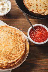 Traditional Russian Crepes Blini stacked in plate and cast-iron frying pan with red caviar, fresh sour cream on dark wooden table. Russian festival meal Maslenitsa or Shrovetide. Top view.