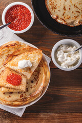 Traditional Russian Crepes Blini stacked in plate and cast-iron frying pan with red caviar, fresh sour cream on dark wooden table. Russian festival meal Maslenitsa or Shrovetide. Top view.