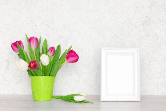 Blank white photo frame with tulip flower bouquet on the table near the wall. Selective focus. Spring mock-up background