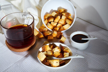 Square toasted pieces of homemade delicious rusk, hardtack, Dryasdust, zwieback, Liquid honey in a saucer and black tee in a cap on a white tablecloth.