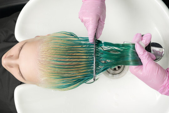 Top view of hairdresser in protective glove holds hair in hand and combs long green and discolored hair of customer, while washing hair in shower in special beauty salon sink.