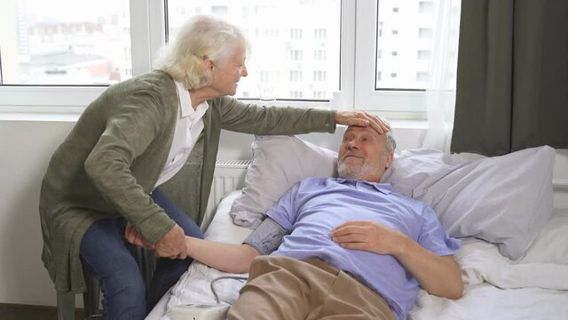 caring elderly woman checks the blood pressure of her old man with hypertension while lying at home in bed, health care.