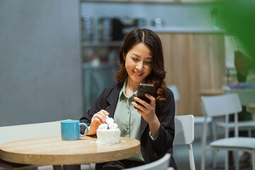 Portrait of young Asian businesswoman drinking coffee, eating cream cake and using phone