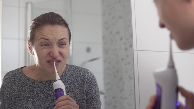 A woman cleans her teeth with an irrigar in the morning in the bathroom, reflected in the mirror.