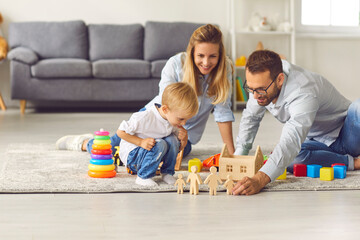 Young parents play with their son wooden figurines sitting on the floor among various toys. Mother,...