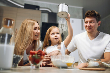 The family is cooking. Daughter, mom and dad are preparing the dough into powder. Live at home in the kitchen. They enjoy and smile. daughter pours flour