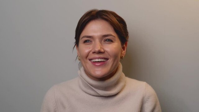Caucasian woman with brown hair is smiling at speaking at the camera during a short video on a gray studio wall