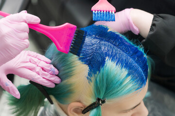 Two hairdressers using pink brush while applying blue paint to female client with emerald hair...
