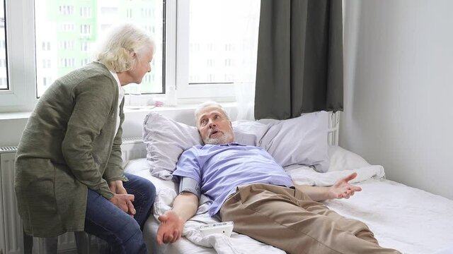 An elderly woman takes care of a man, helps to measure blood pressure with a tonometer. A man is lying on a bed with high blood pressure.