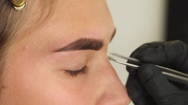Closeup view 4k video of client face and stylist's hands working in gloves. Master removes hair using tweezers while making correct trendy beautiful shape of female brows