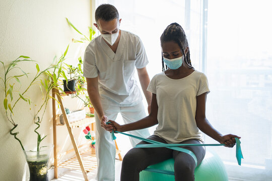 African Woman With Mask Doing Rehabilitation Exercises On Her Legs With An Elastic Band And A Medicine Ball