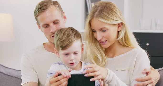 Smiling young American family sit on couch with little kid watch cartoons on tablet, happy caucasian parents have fun relax with boy child on sofa enjoy video on pad spending time at home together.