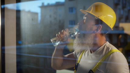View through window of aged construction worker drinking water on location site.