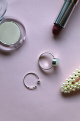 Silver rings, pearl barrette and various make up products on pale pink background. Flat lay.