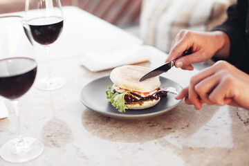 Eating burger in a restaurant with fork and knife. Two glasses of red wine on a marble table