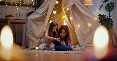 Mother and little daughter with tablet computer playing in teepee at home in evening