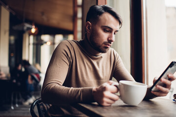 Thoughtful young man using smartphone and drinking coffee in cafeteria