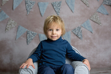 Naklejka premium Cute blond toddler child, boy, sitting on little baby armchair, reading a book and holding flowers