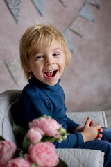 Fototapeta premium Cute blond toddler child, boy, sitting on little baby armchair, reading a book and holding flowers
