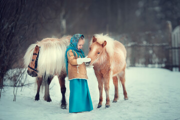 Russian style. Beautiful little girl in blue pavloposad shawl is feeding pony in winter in Russia. Image with selective focus and toning