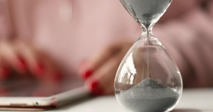 Hourglass black sand in a transparent flask. Female hands holding a tablet on white table background.
