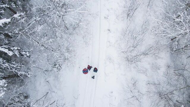 family of three stands in a snow covered forest in winter and waves to a drone that soars up into the sky